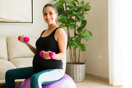 Cheerful beautiful woman with a big belly enjoying her pregnancy and smiling while working out with a fitness ball and dumbbellsの写真素材