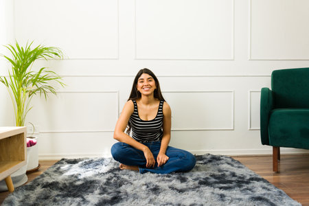 Smiling Hispanic woman sitting cross-legged on a rug in a bright living roomの写真素材