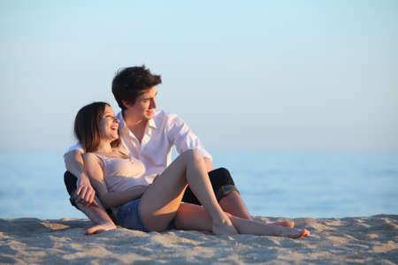 Couple sitting and laughing on the sand of the beach at sunset with the sea in the backgroundの写真素材