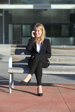 Businesswoman talking on the mobile phone sitting on a bench in front the officeの写真素材