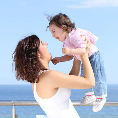 Pretty mum laughing and raising her baby girl with the sea in the backgroundの写真素材