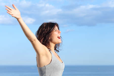 Attractive woman with raised arms shouting to the wind in the beach with the sea and blue sky in the backgroundの写真素材