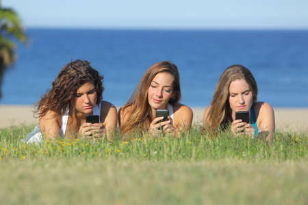 Group of three teenager girls typing on the mobile phone lying on the grass with the sea in the backgroundの写真素材