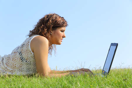 Profile of a beautiful woman lying on the grass browsing a laptop with a blue sky in the background              の写真素材