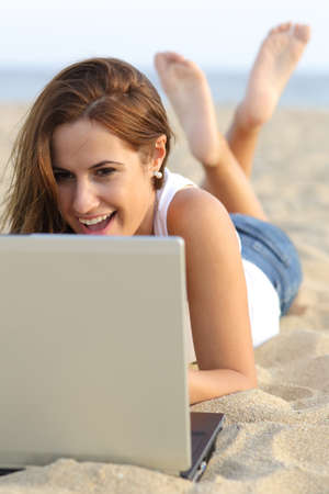 Front view of a teenager girl laughing browsing her laptop lying on the sand of the beachの写真素材