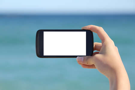 Woman hand showing a blank smart phone horizontal screen display on the beach with the sea in the...