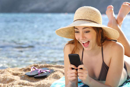 Funny surprised woman watching social media in a smart phone on the beach on vacations                の写真素材