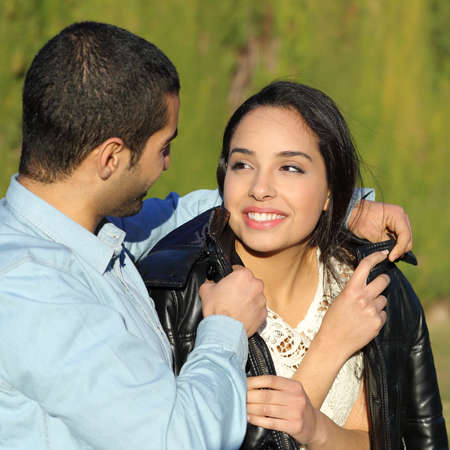 Happy arab couple flirting while gentleman cover her with his jacket in a park with a green background               の写真素材