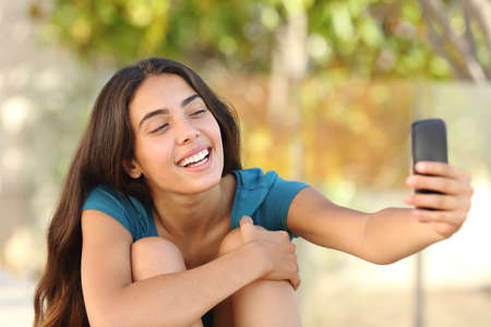 Happy laughing teen girl taking a selfie portrait with her smart phone in a parkの写真素材