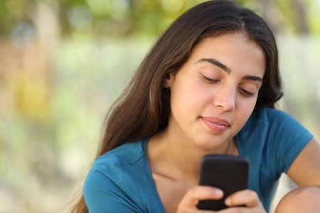 Pretty happy teenager girl texting in a smart phone in a park with a soft green backgroundの写真素材