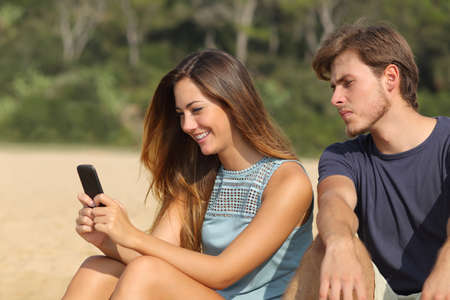 Jealous boyfriend watching his girlfriend texting on the phone on the beachの写真素材