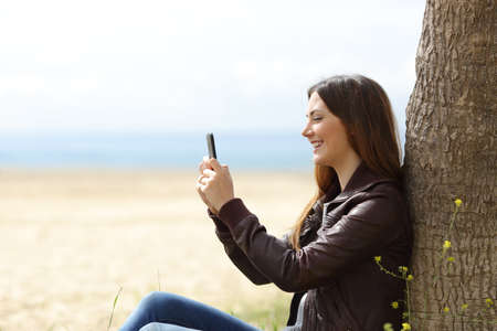 Happy woman texting in the phone sitting on the beach in winter with the sea in the background