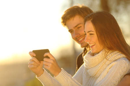Happy couple watching media videos in a smart phone at sunset on the beachの写真素材