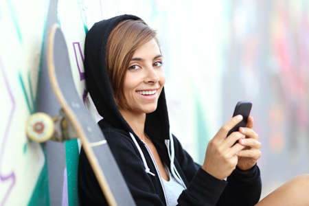 Teenager skater girl using a smart phone looking at camera leaning in a wall with graffitiの写真素材