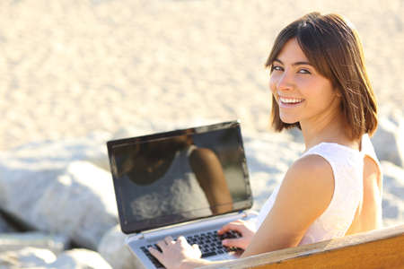 Happy woman typing on a laptop and looking at camera sitting on a benchの写真素材
