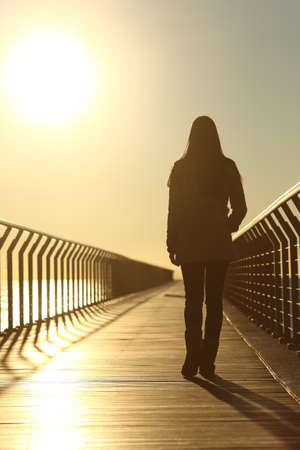 Sad woman silhouette walking alone on a bridge on the beach in winter at sunsetの写真素材
