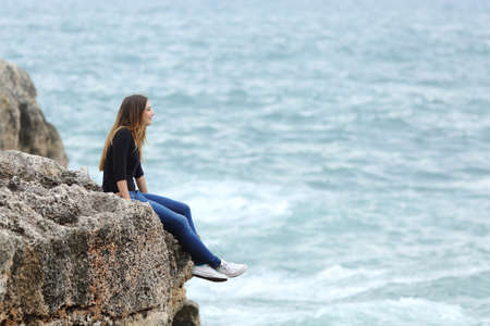 Side view of a full body of a casual woman thinking sitting in a cliff watching the seaの写真素材