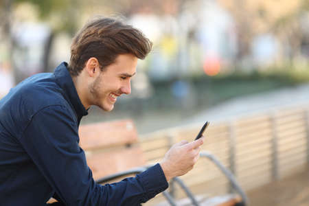 Profile of a happy guy using a smart phone sitting on a bench in a parkの写真素材