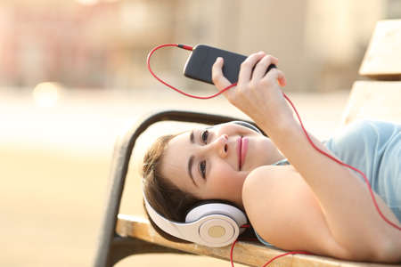 Teen girl listening to the music from a smart phone lying in a bench of a parkの写真素材