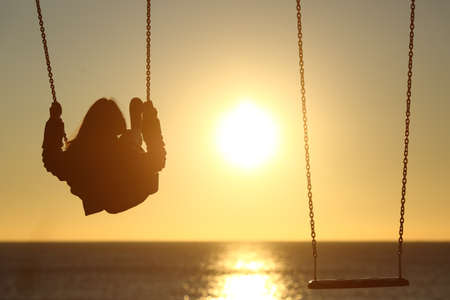 Back light of a lonely woman silhouette swinging at sunset on the beach with another empty swingの写真素材