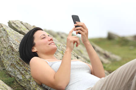 Happy woman using a mobile phone lying in the mountain with a white isolated sky in the backgroundの写真素材