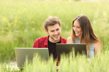 Couple or friends watching apps in two laptops in a green fieldの写真素材