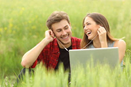 Happy couple or friends sharing music from a laptop sitting in a green field and looking each otherの写真素材