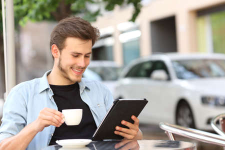 Happy man reading an ebook or tablet in a coffee shop terrace holding a cup of teaの写真素材