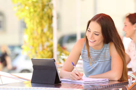 Happy student studying and learning taking notes with a digital tablet in a coffee shop