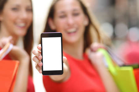 Two joyful shoppers with shopping bags showing a blank smart phone screen in the street