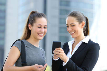Two businesswomen talking about smart phone in the street with office buildings in the backgroundの写真素材