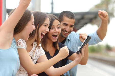 Four euphoric multi ethnic caucasian and arab friends watching tv on a a tablet in a train station