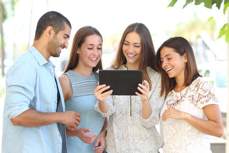 Front view of a group of four happy friends watching videos on a tablet in the streetの写真素材