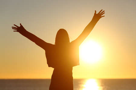 Free happy woman raising arms watching the sun in the background at sunrise Free happy woman raising arms watching the sun in the background at sunrise