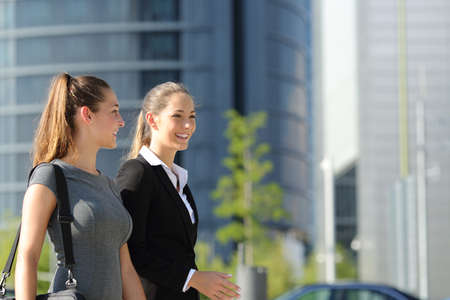 Two happy businesswomen walking and talking in the street with office buildings in the backgroundの写真素材