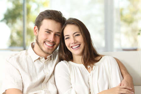 Happy couple sitting on a couch at home and looking at cameraの写真素材
