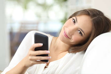 Beautiful woman using a smart phone sitting on a comfortable couch at home and looking at cameraの写真素材