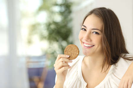 Happy girl showing a dietetic cookie sitting on a couch at homeの写真素材