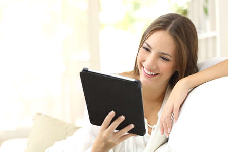 Happy woman reading an ebook or tablet sitting on a couch at homeの写真素材