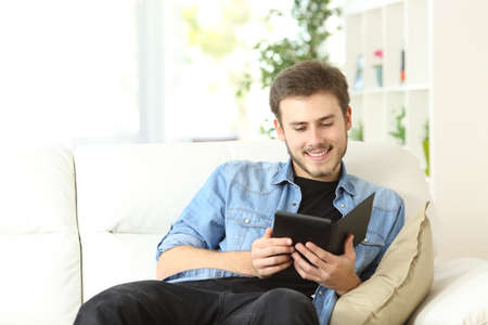 Happy man reading a book in an ebook reader sitting on a couch at homeの写真素材