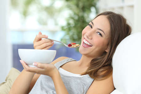Happy girl eating cereals with fruits from a bowl sitting on a couch at homeの写真素材
