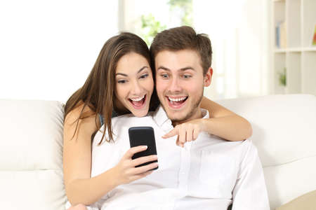 Attractive surprised and amazed couple watching smart phone together sitting on a couch in a living room at home