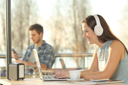 Side view of a beautiful female working on line with headphones typing in a laptop in a restaurant indoor with a window in the background and people outdoorsの写真素材