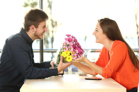 Profile of a couple dating and looking each other with a man giving a bouquet of flowers to his partner in a coffee shopの写真素材
