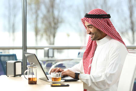 Arab saudi man working online with a laptop and smartwatch in a coffee shop or an hotel bar with a...