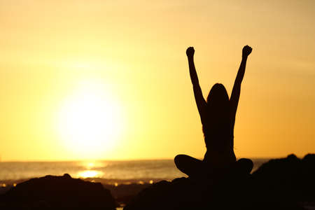 Back view of an excited euphoric woman silhouette raising arms and looking orange sun at sunrise in the beach with the ocean in the backgroundの写真素材