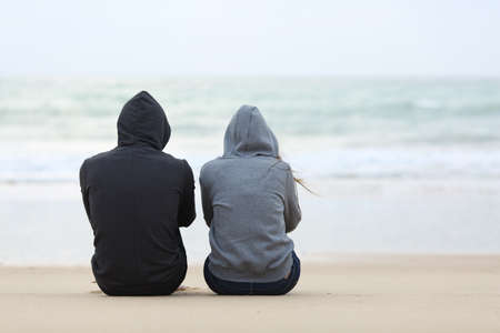 Back view of two sad teenagers sitting on the sand of the beach and looking at horizon in a bad weather dayの写真素材