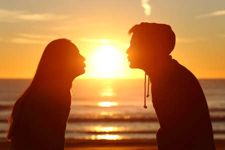 Side view of a couple silhouette of teenagers kissing the sun with love at sunset on the beach with the horizon in the backgroundの写真素材