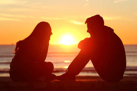 Side view of full body of two friends or couple silhouette of teens facing at sunset on the beach with the sun in the middleの写真素材