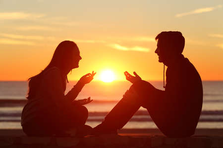 Side view of a full body of two friends or couple silhouette of teens sitting and talking at sunrise on the beach with the sun in the middleの写真素材
